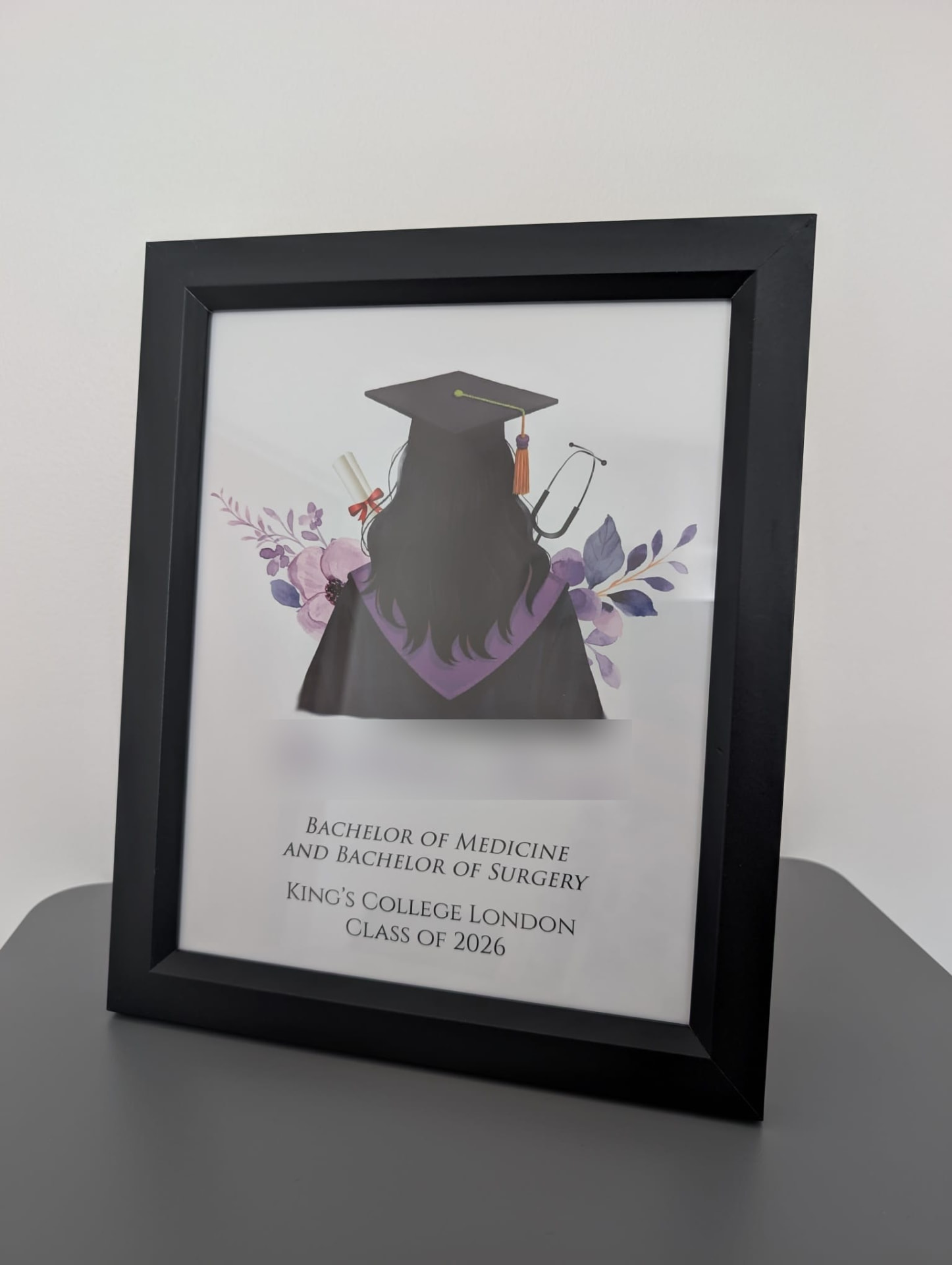 Black-framed graduation print: an illustrated graduate seen from behind in cap and gown with a stethoscope and scroll, above the degree, university and year