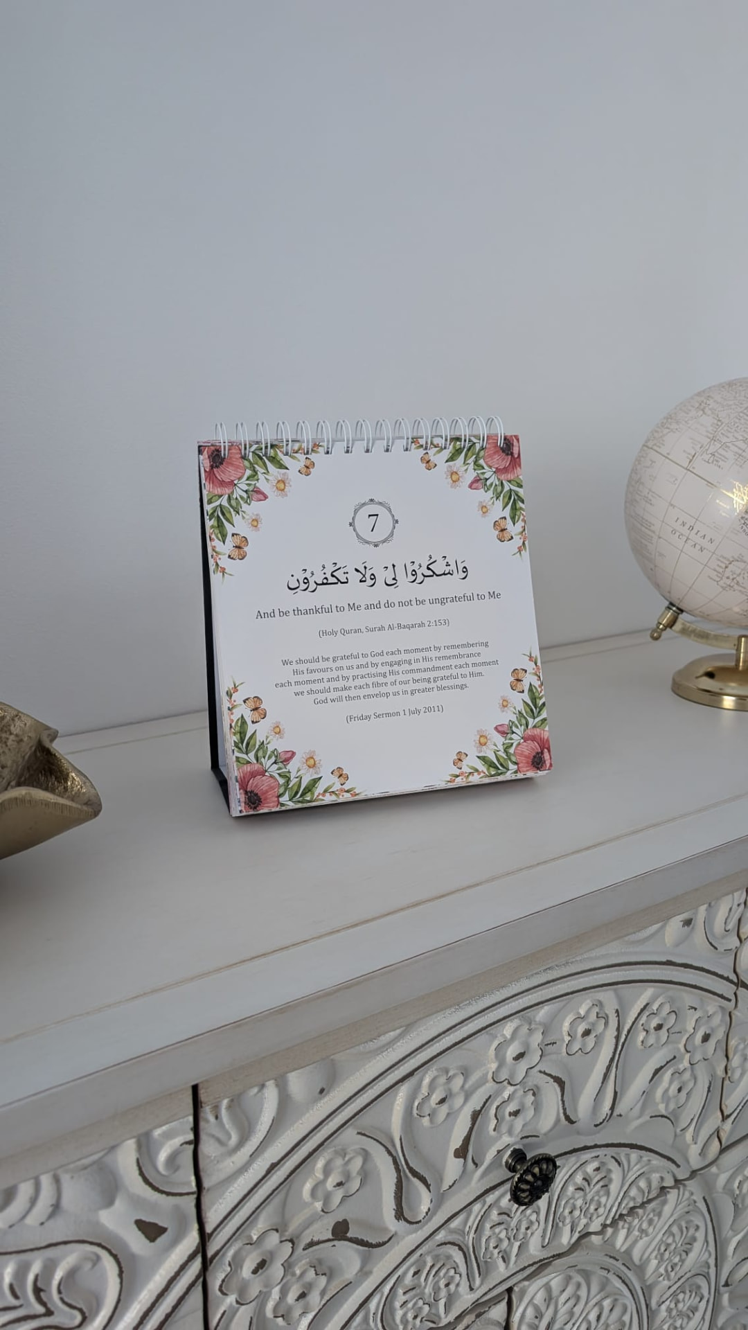 Spiral-bound floral desk calendar standing on a shelf, open at day 7 with a Quran verse, its translation and a short reflection