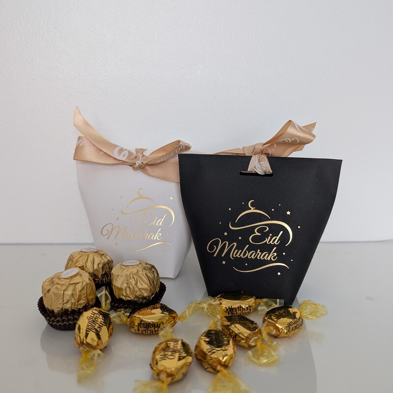White and black Eid Mubarak gift bags with gold foil script and ribbon, beside chocolates