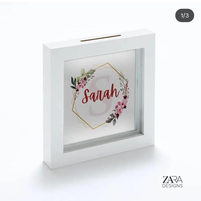 White box-frame money box printed with a floral wreath, a gold geometric frame and the name Sarah in red script